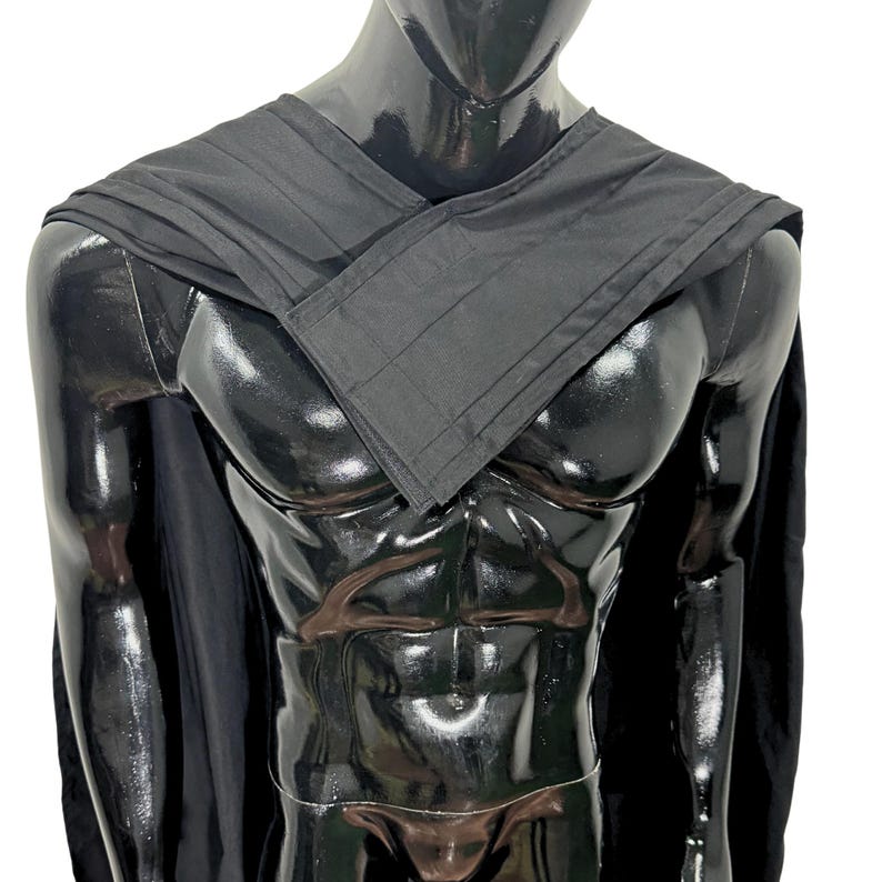 May include: A black cape featuring a structured, layered collar design that wraps across the chest. The garment is made of a solid black fabric with visible stitching along the edges of the collar and shoulder panels, draping over the shoulders.