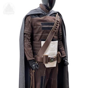 May include: A brown long-sleeved tunic and trousers set featuring white rectangular panels, black accents, and white stitching. A dark gray cape drapes over the shoulders, accompanied by a brown utility belt with pouches and a diagonal bandolier.