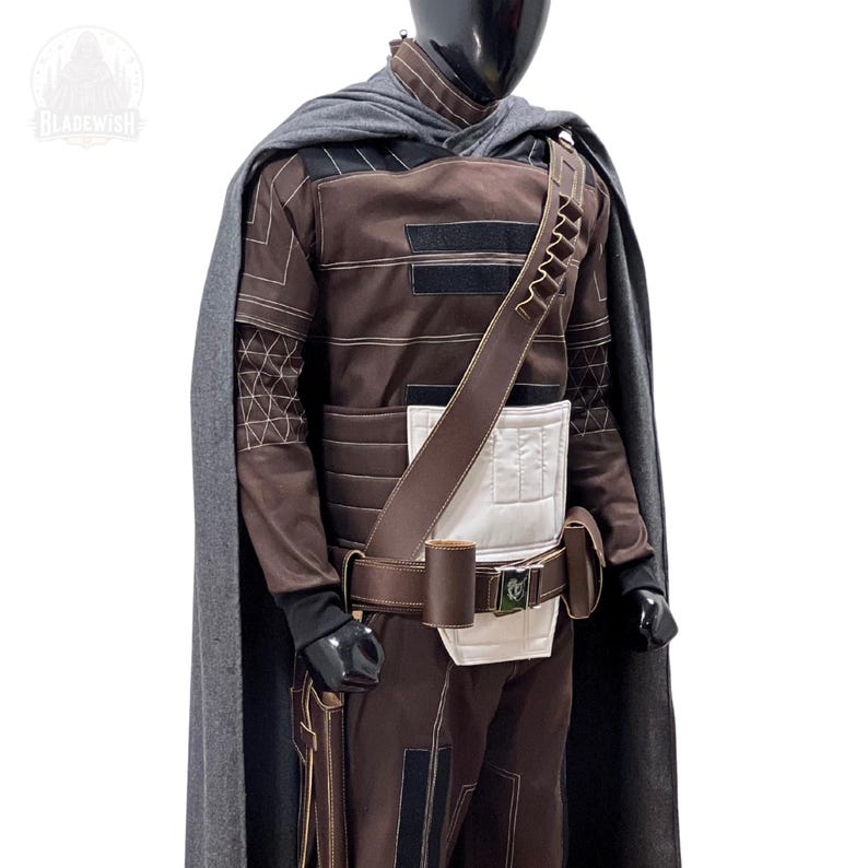 May include: A brown long-sleeved tunic and trousers set featuring white rectangular panels, black accents, and white stitching. A dark gray cape drapes over the shoulders, accompanied by a brown utility belt with pouches and a diagonal bandolier.