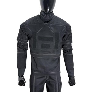 May include: A black tactical-style long-sleeve shirt and matching trousers set featuring a high collar, padded shoulder panels, and prominent white contrast stitching. The shirt includes rectangular hook-and-loop fastener patches on the chest.