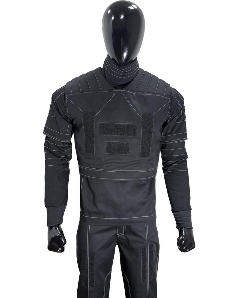 May include: A black tactical-style long-sleeve shirt and matching trousers set featuring a high collar, padded shoulder panels, and prominent white contrast stitching. The shirt includes rectangular hook-and-loop fastener patches on the chest.