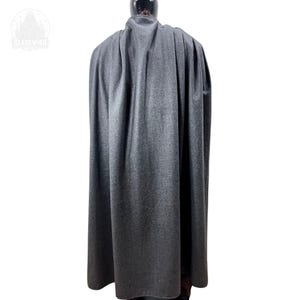 May include: A floor-length charcoal gray cloak featuring a high collar and vertical pleats that drape down the back. The garment is displayed on a mannequin, showcasing a textured fabric surface and a consistent dark gray color throughout the piece.