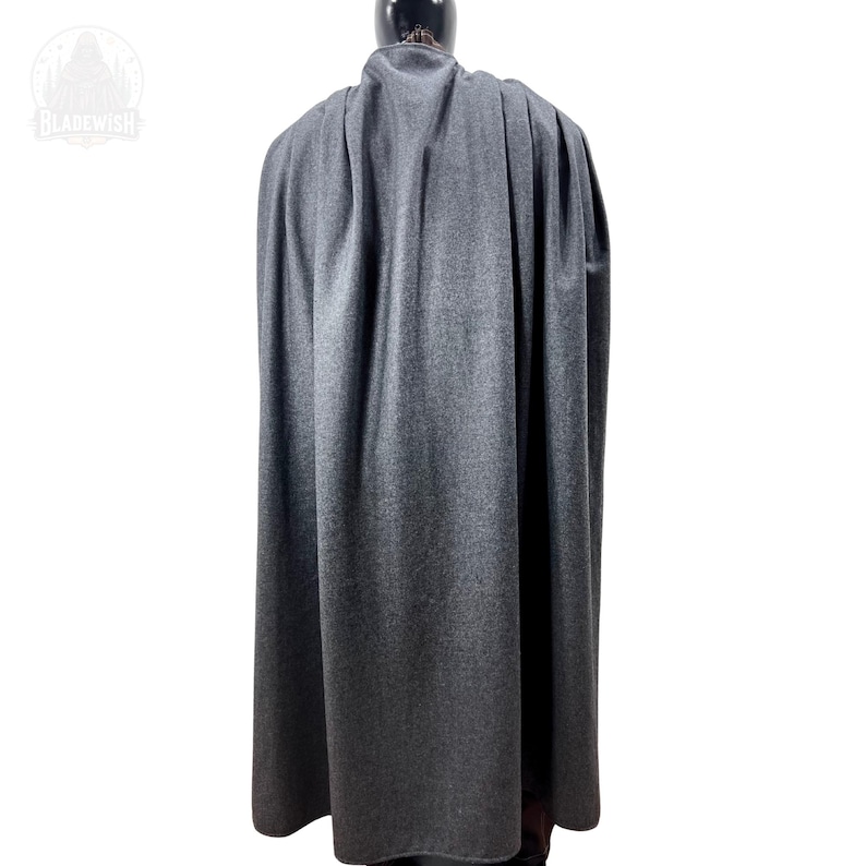 May include: A floor-length charcoal gray cloak featuring a high collar and vertical pleats that drape down the back. The garment is displayed on a mannequin, showcasing a textured fabric surface and a consistent dark gray color throughout the piece.