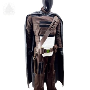 May include: A brown costume set featuring a long grey cape, a brown tunic with white chest panel, and matching trousers. The outfit includes a brown utility belt with a green buckle, a shoulder strap with ammunition loops, and a small pouch.