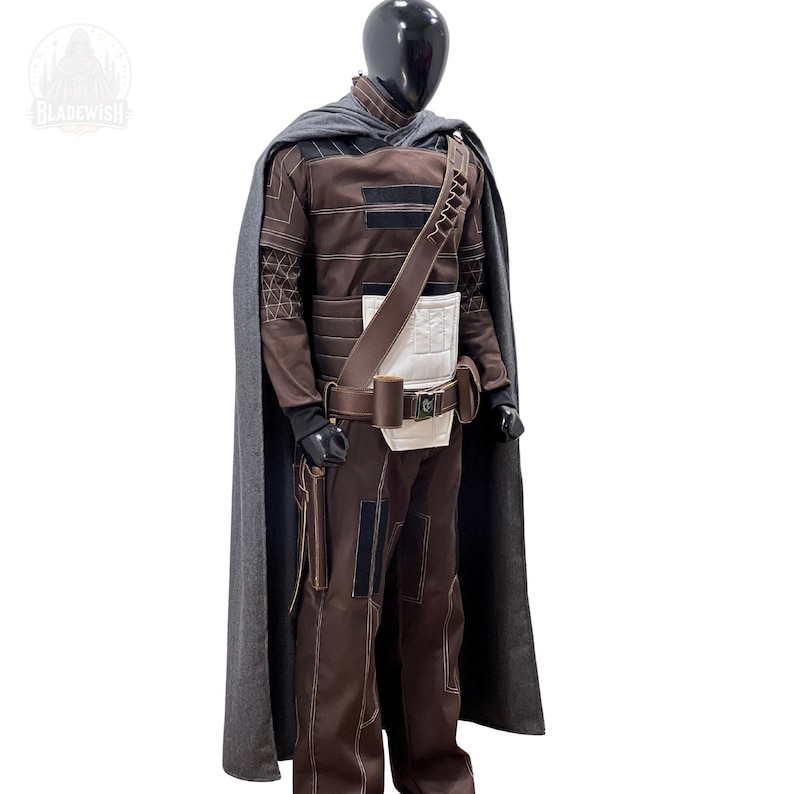 May include: A brown and dark brown costume set featuring a long grey cape, a structured jacket with white paneling, and matching trousers. The outfit includes a brown utility belt with pouches, a diagonal shoulder strap, and visible stitching details.