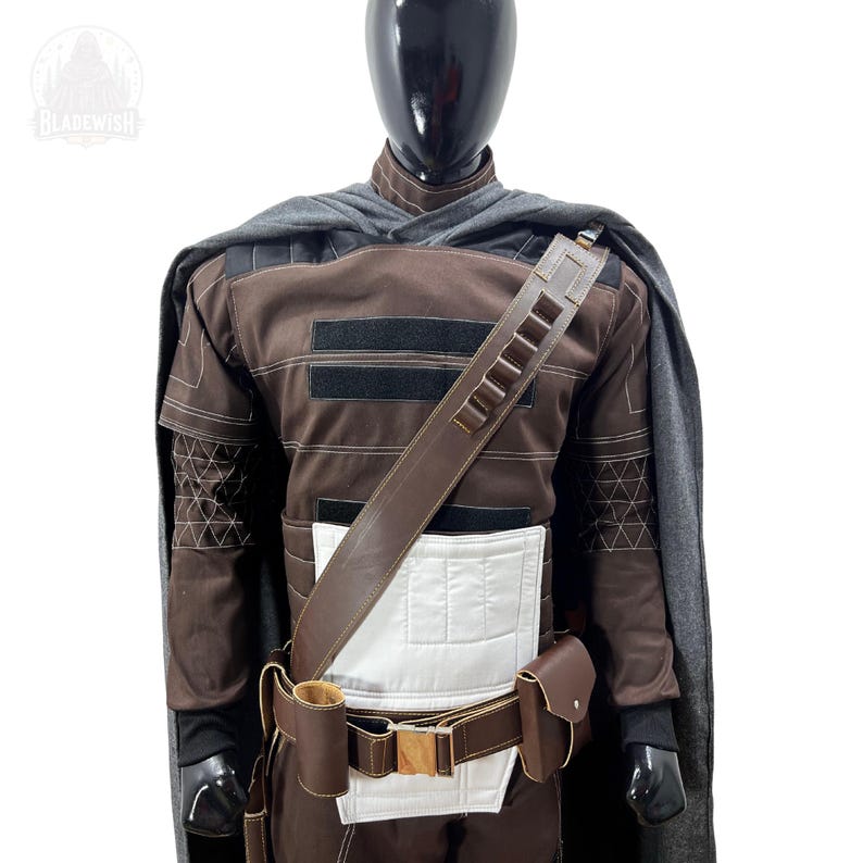 May include: A brown and black tactical costume featuring a grey cape, a white chest panel, and a brown utility belt with pouches and a bandolier. The garment includes black velcro strips on the chest and geometric stitching on the sleeves.
