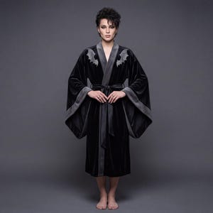 May include: Black velvet robe with gray trim and bat embroidery on the lapel. The robe has wide sleeves and a matching belt. The model is standing in front of a gray background.