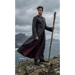 May include: A long, black coat with a burgundy lining, worn by a person standing on a rocky cliffside. The coat is buttoned and flows open at the front. The person holds a wooden staff. The background features a cloudy sky and ocean.