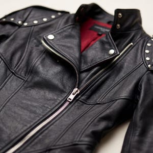 K&ouml;nnte beinhalten: Schwarze Lederjacke mit silberfarbenen Rei&szlig;verschl&uuml;ssen und Nieten an den Schultern. Die Jacke hat ein rotes Innenfutter und einen klassischen Biker-Stil.