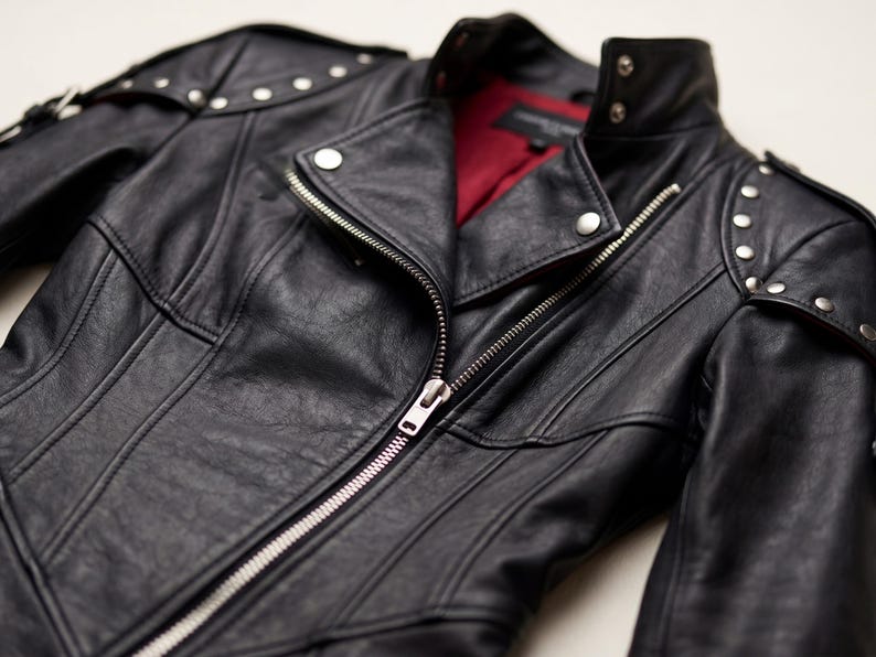 K&ouml;nnte beinhalten: Schwarze Lederjacke mit silberfarbenen Rei&szlig;verschl&uuml;ssen und Nieten an den Schultern. Die Jacke hat ein rotes Innenfutter und einen klassischen Biker-Stil.