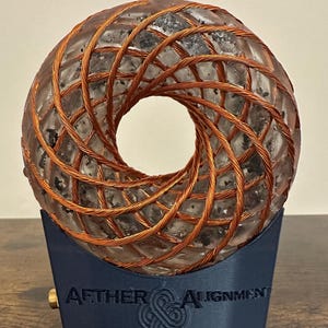 May include: A circular, translucent resin object embedded with dark flecks and wrapped in a copper wire coil. It rests on a dark blue, trapezoidal base featuring the embossed text AETHER ALIGNMENT and a central knot-like emblem.