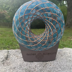 May include: A circular, translucent blue resin sculpture featuring an intricate, spiraling copper wire inlay. The sculpture is mounted on a dark brown, trapezoidal base with the embossed text Aethet Alignment and a central knot symbol.