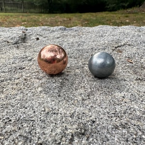 Puede incluir: Dos objetos esféricos sobre una superficie texturizada. Una esfera es de cobre pulido con un acabado metálico de color marrón rojizo cálido, mientras que la otra esfera es de acero cepillado con un aspecto metálico gris mate y frío.