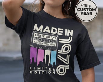 May include: Black t-shirt with a retro design. The shirt features the text "MADE IN '76" with a cassette tape graphic and the words "BEST OF '76" and "LEGENDARY HITS." Below the tape is a skyline graphic with purple, pink, and blue stripes. The words "LIMITED EDITION" are also printed on the shirt.