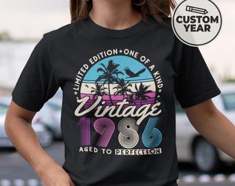 May include: A black short-sleeve t-shirt featuring a graphic print with palm trees, a bird, and the text: LIMITED EDITION ONE OF A KIND, Vintage 1986, AGED TO PERFECTION. A circular badge in the upper right corner displays a pencil icon and CUSTOM YEAR.