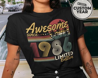 May include: Black t-shirt with the text "Awesome since 1986 Limited Edition" in yellow, red, and green. The design includes a sunset graphic with palm trees. A round sticker on the upper right says "Custom Year".