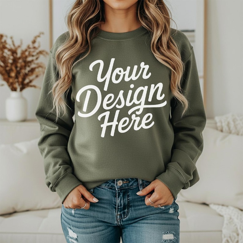 May include: An olive green sweatshirt with the text "Your Design Here" in white script. The sweatshirt has long sleeves and a crew neck. The model is wearing blue jeans.