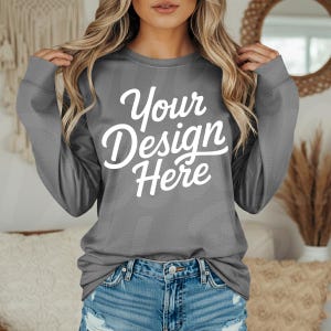 May include: A gray long-sleeve sweatshirt with the text "Your Design Here" in white script. The sweatshirt is paired with denim shorts. The image is a product mockup, ideal for showcasing custom designs.