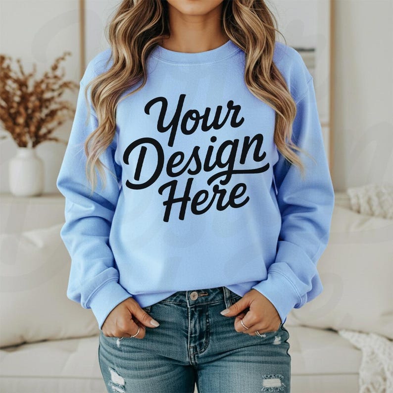 May include: A light blue sweatshirt with the text "Your Design Here" in black script. The sweatshirt has long sleeves and a crew neck. The model is wearing blue jeans.