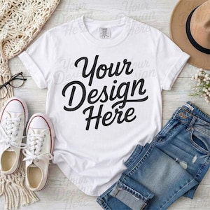 May include: A white t-shirt with the text "Your Design Here" in black script. The shirt is paired with blue jeans, white sneakers, and a brown hat. The image is styled on a light wood surface.