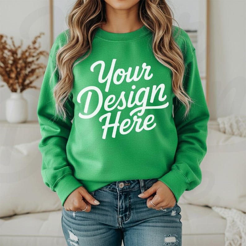 May include: A green sweatshirt with the text "Your Design Here" in white script. The sweatshirt is a solid green color and has long sleeves. The model is wearing blue jeans.