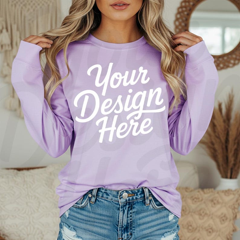 May include: A long-sleeved, light purple sweatshirt with the text "Your Design Here" in white script. The shirt is a solid color and has a crew neck. The model is wearing denim shorts.