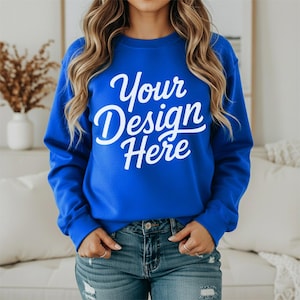 May include: A royal blue sweatshirt with the text "Your Design Here" in white script. The sweatshirt is being worn with blue jeans. The model is wearing rings on their fingers.
