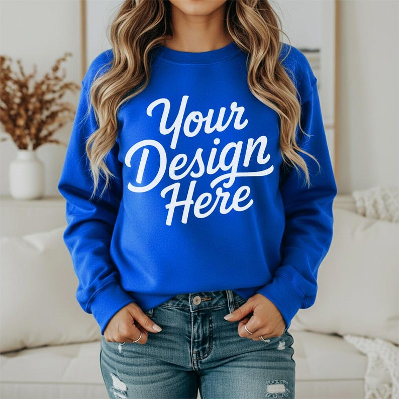 May include: A royal blue sweatshirt with the text "Your Design Here" in white script. The sweatshirt is being worn with blue jeans. The model is wearing rings on their fingers.