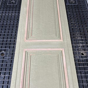 Puede incluir: Un panel de puerta rectangular verde salvia con molduras empotradas de color marr&oacute;n claro. El panel presenta dos secciones rectangulares, cada una con una serie de marcos anidados. El panel de la puerta est&aacute; sobre una superficie negra con patr&oacute;n de rejilla.