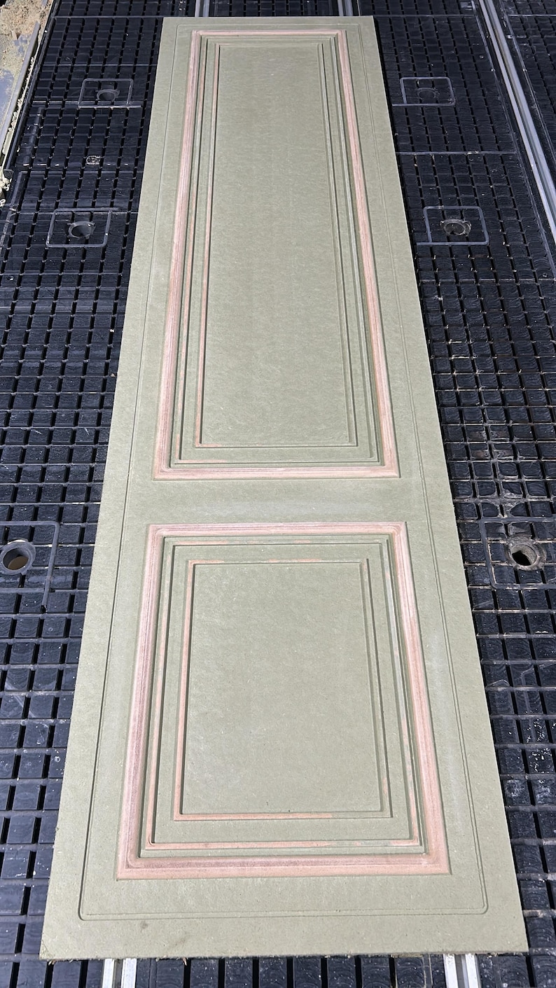Puede incluir: Un panel de puerta rectangular verde salvia con molduras empotradas de color marr&oacute;n claro. El panel presenta dos secciones rectangulares, cada una con una serie de marcos anidados. El panel de la puerta est&aacute; sobre una superficie negra con patr&oacute;n de rejilla.
