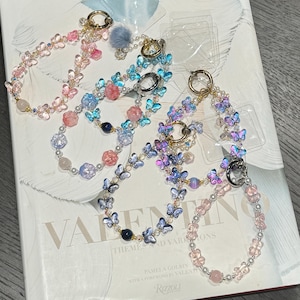 Puede incluir: Cinco pulseras de amuleto de mariposa de colores con cuentas y amuletos de diferentes colores. Las pulseras están hechas con cuentas rosas, azules y moradas y amuletos de mariposa. Las pulseras están sobre una superficie blanca con un libro en el fondo.