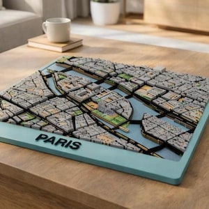 Puede incluir: Un mapa 3D de París, Francia, con un borde azul claro. El mapa presenta representaciones detalladas de edificios, calles y el río Sena. La palabra "PARIS" se muestra en letras negras en el borde.