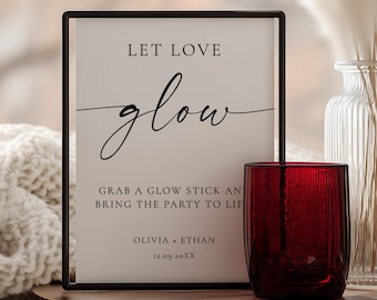 May include: A framed sign with the words "LET LOVE glow" in elegant script, next to a red glass votive candle holder. The sign also includes the text "GRAB A GLOW STICK AND BRING THE PARTY TO LIFE" and the names "OLIVIA + ETHAN" with a date.