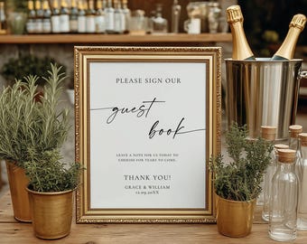 May include: A framed sign with gold trim reads "PLEASE SIGN OUR guest book" in elegant script. The sign is on a wooden table with potted plants, champagne bottles, and clear glass bottles. The sign also says "LEAVE A NOTE FOR US TODAY TO CHERISH FOR YEARS TO COME" and "THANK YOU! GRACE & WILLIAM 12.09.20XX".