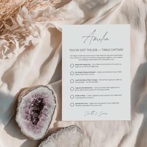 Puede incluir: Una tarjeta rectangular blanca con texto negro titulada Amelia, You've Got The Job - Table Captain, que enumera seis tareas para los invitados a la boda. La tarjeta está colocada junto a dos geodas de amatista y cuarzo en bruto sobre una tela crema.