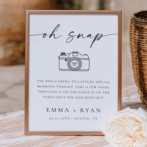 May include: A framed sign with the words "oh snap" in elegant script above a line drawing of a camera with a heart. The sign includes instructions for capturing special moments and the names Emma and Ryan, along with a date and location.