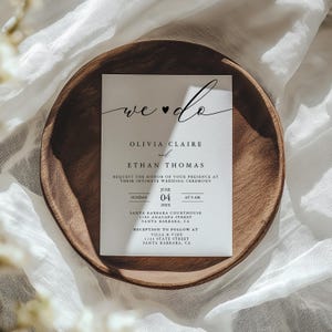 May include: A white wedding invitation with black calligraphy that reads "we ♥ do" followed by the names Olivia Claire and Ethan Thomas. The invitation details the wedding ceremony and reception to be held on Sunday, June 4th, 20XX at the Santa Barbara Courthouse and Villa & Vine.