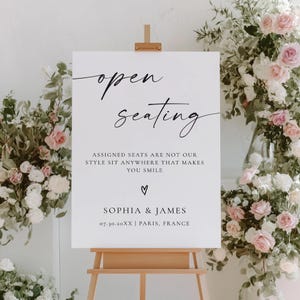 Puede incluir: Un letrero blanco sobre un caballete de madera con las palabras "open seating" en escritura elegante. El letrero también incluye el texto "ASSIGNED SEATS ARE NOT OUR STYLE SIT ANYWHERE THAT MAKES YOU SMILE" y los nombres "SOPHIA & JAMES". Arreglos florales enmarcan el letrero.