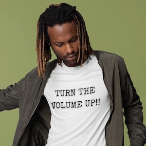 May include: White t-shirt with the text "TURN THE VOLUME UP!!" in black. The shirt is worn with a dark green bomber jacket. The background is a solid green color.
