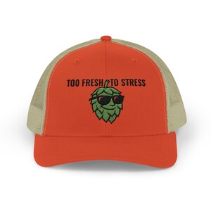 May include: A trucker hat with a red front panel and a tan mesh back. The hat features the text "TOO FRESH TO STRESS" above a green hop wearing sunglasses.