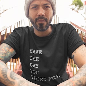 Peut inclure: Une personne portant un t-shirt noir avec le texte "HAVE THE DAY YOU VOTED FOR." imprimé en blanc.