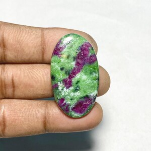May include: An oval-shaped cabochon gemstone featuring a vibrant mix of bright green zoisite and deep magenta-colored ruby inclusions. The polished stone displays a smooth, glossy surface with natural color variations and dark mineral speckles.