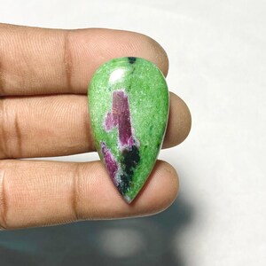 May include: A pear-shaped cabochon gemstone featuring a vibrant green zoisite base with prominent inclusions of deep magenta-colored ruby and small patches of black mineral deposits, polished to a smooth, reflective finish with a rounded top.