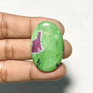 May include: An oval-shaped cabochon gemstone featuring a vibrant lime green base color with a distinct, irregular magenta-colored inclusion on the left side and small dark black speckles scattered across the lower portion of the polished surface.