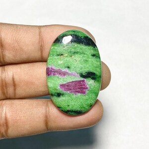 May include: An oval-shaped cabochon gemstone featuring a vibrant green zoisite base with distinct, opaque magenta-colored ruby inclusions and dark black hornblende mineral streaks distributed across the polished, smooth surface of the stone.