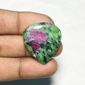 May include: A teardrop-shaped zoisite gemstone featuring a vibrant green base with prominent inclusions of deep magenta ruby and scattered black mineral flecks. The stone has a smooth, polished surface with a reflective sheen across the top.