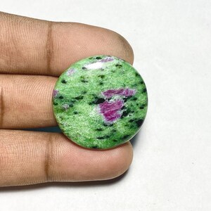 May include: A round, polished cabochon gemstone featuring a vibrant green zoisite base with prominent, irregular magenta-colored ruby inclusions and scattered black mineral flecks throughout the smooth, glossy surface of the stone.