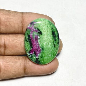 May include: An oval-shaped cabochon gemstone featuring a vibrant green zoisite base with prominent inclusions of deep magenta-colored ruby crystals and scattered black mineral patches, polished to a smooth, glossy finish with a rounded surface.