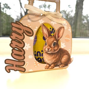 May include: A decorative Easter gift box in light brown, with a handle and a cream ribbon. It features a cutout with a brown bunny illustration and a chocolate egg. The name "Harry" is written on the side.