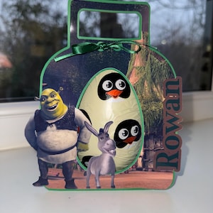 May include: A green gift bag with a handle, decorated with Shrek, Donkey, and an egg with penguin faces. The bag has a green bow and the name "Rowan". The background shows a forest scene.